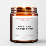 a burning candle in a brown amber jar. The name of the candle in black bold font is ADHD Small Business Owner. The scent is Cashmere. Red Virgins On Fire Logo.