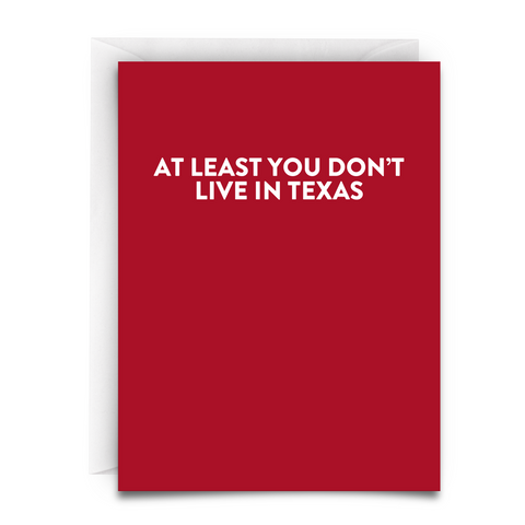 A greeting card. Red. White font. Says, "At Least You Don't Live in Texas." White Envelope.