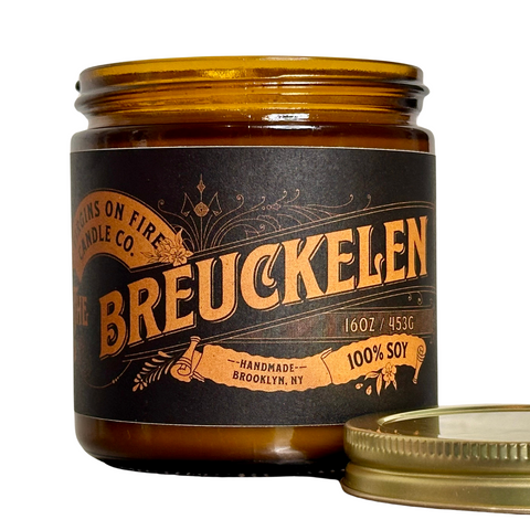 Hand-poured 16oz amber glass jar candle labeled 'Breuckelen' with vintage-inspired black and gold typography, made with 100% soy wax by Virgins on Fire Candle Co. in Brooklyn, NY