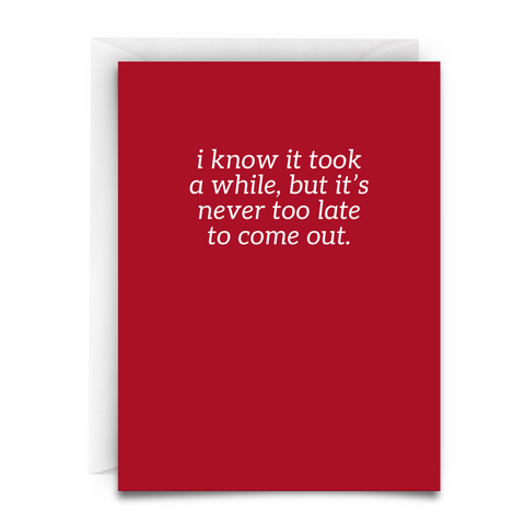 A greeting card against a white envelope background. The card has a deep red background with white text in an italic serif font that reads 'i know it took a while, but it's never too late to come out.' The message offers support and validation for those coming out as LGBTQ+